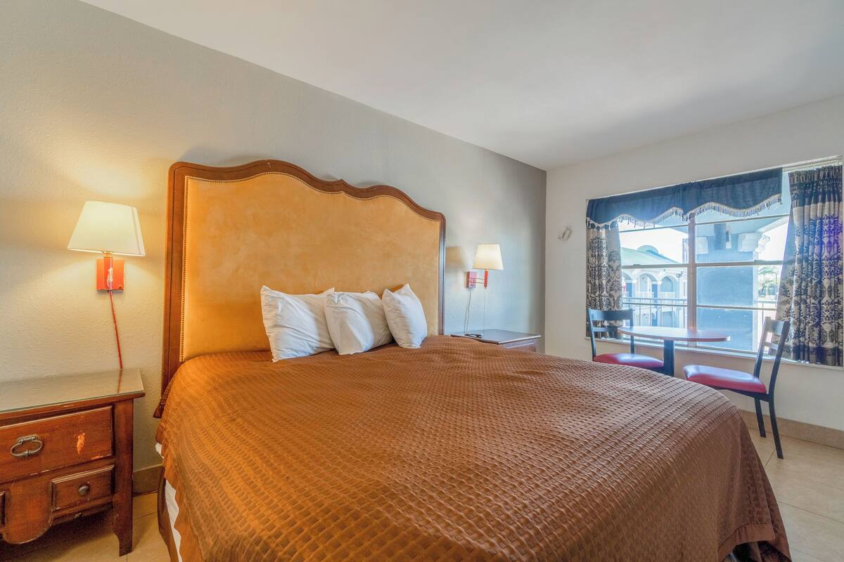 A hotel room with a large bed, two bedside tables with lamps, a small table and chair by the window, and a view of buildings outside.
