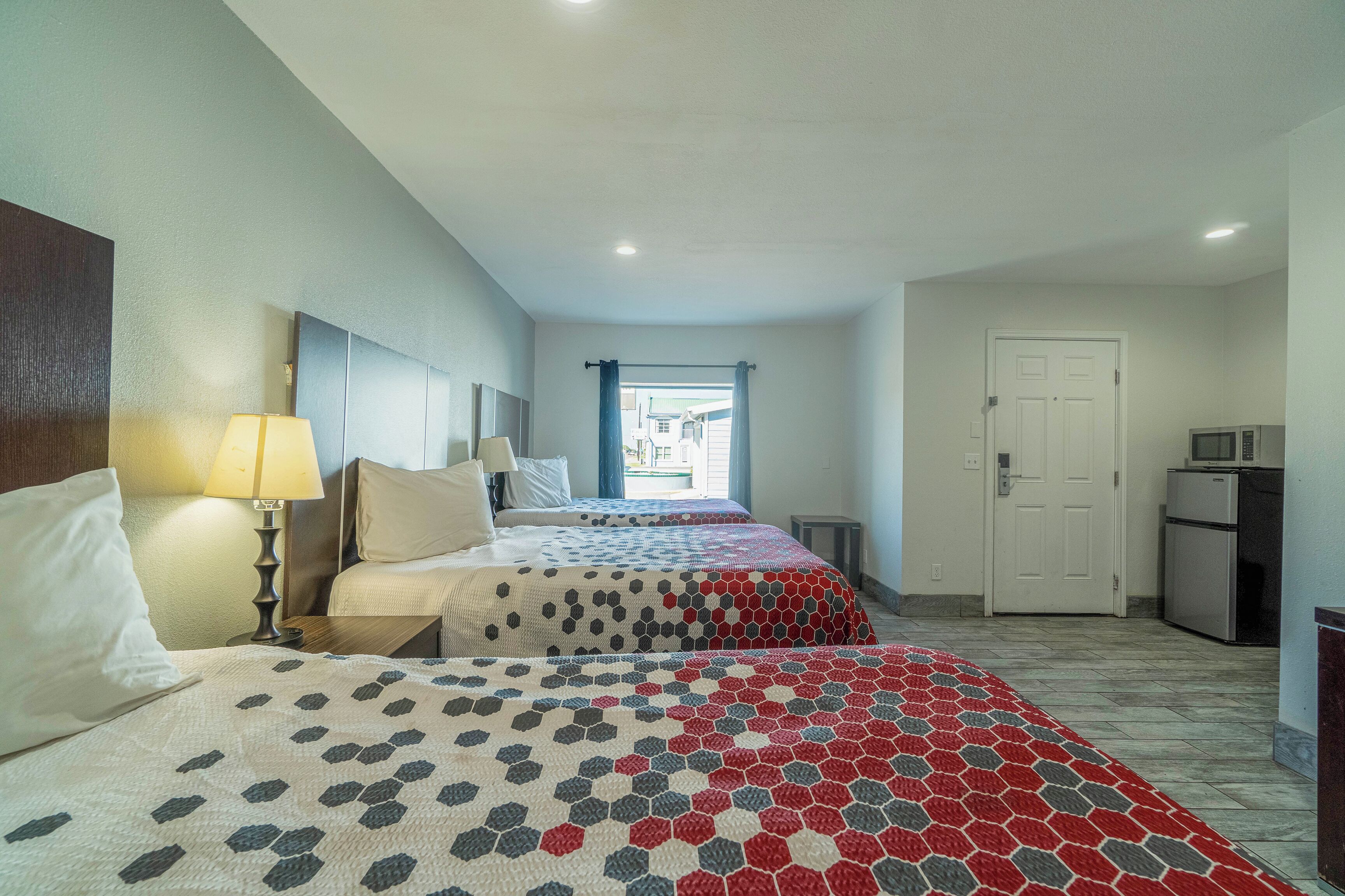 A hotel room with two beds, a desk, a lamp, a microwave, and a door leading to the kitchen.