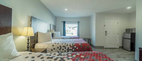 A hotel room with two beds, a desk, a lamp, a microwave, and a door leading to the kitchen.
