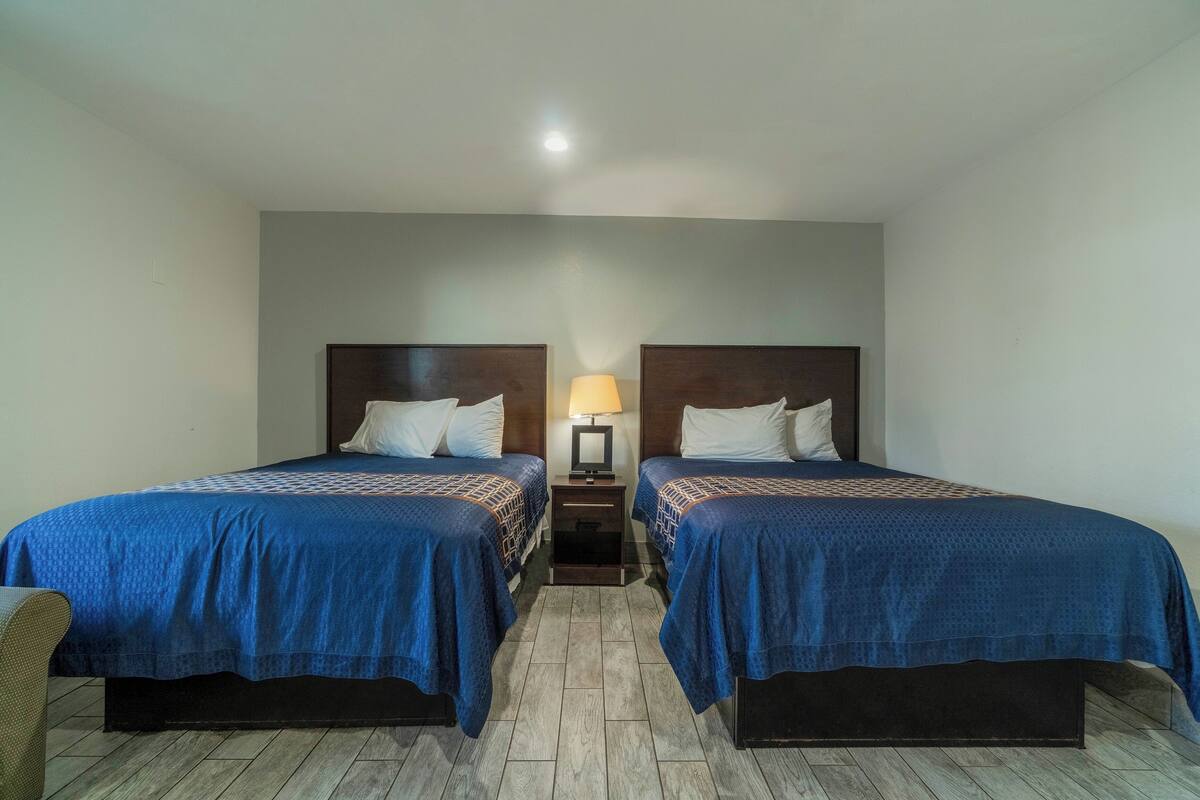 A hotel room with two beds, a nightstand, a lamp, and a chair.