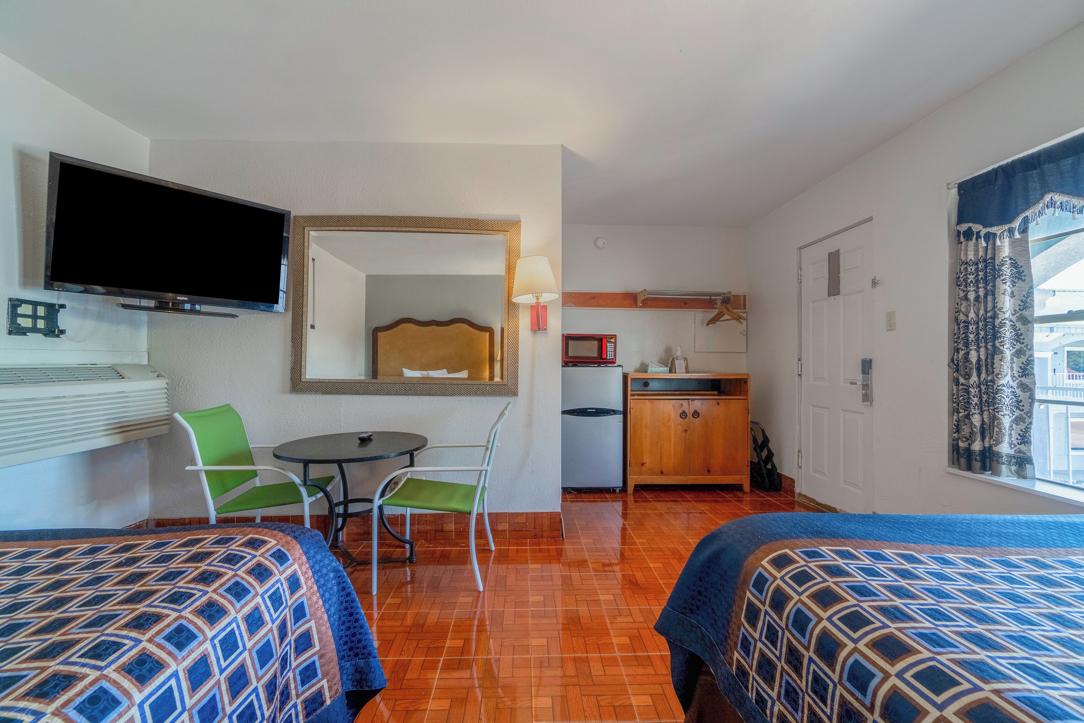 Une chambre d’hôtel avec deux lits, une télévision, une petite table avec des chaises, un coin cuisine et une fenêtre avec des rideaux.