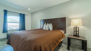 A hotel room with a large bed, a bedside table with a lamp, a window with curtains, and a view of a building outside.