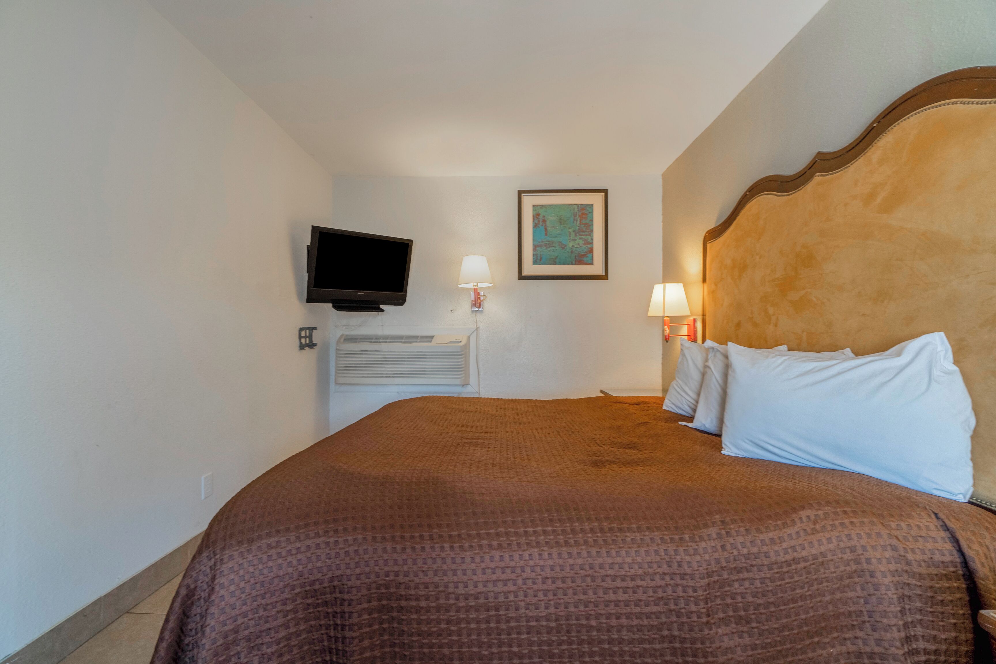 A hotel room with a large bed, a flat-screen TV mounted on the wall, a framed artwork above the bed, and a bedside lamp.