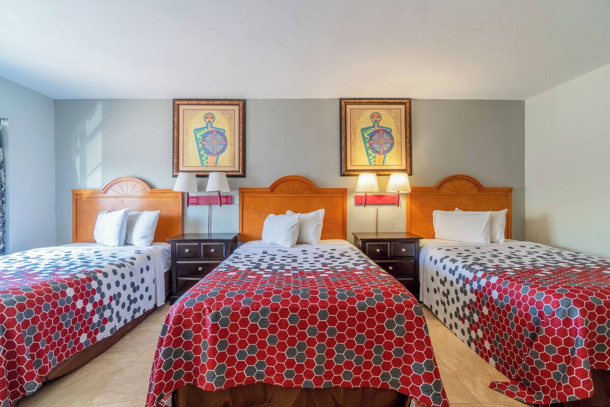 A hotel room with three beds, each with a patterned bedspread, wooden headboards, bedside lamps, and two wall-mounted paintings.