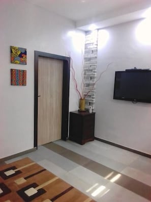A modern living room with a wooden door, a flat-screen TV mounted on the wall, a decorative vase with branches, and a patterned rug. - Premiership UK Suites (Lagos)