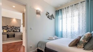 A bedroom with a bed, a nightstand, a wall-mounted lamp, and a view of a kitchen through an open door.