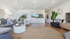 A spacious living area with a modern sofa, a round coffee table, and a dark blue ottoman. There is a kitchen in the background with white cabinets and a large window. - Oceanview Kiama Luxury Coastal Accommodation (Kiama)