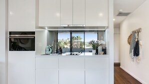 A modern kitchen with white cabinetry, a built-in oven, and a view of palm trees through large windows. - Oceanview Kiama Luxury Coastal Accommodation (Kiama)