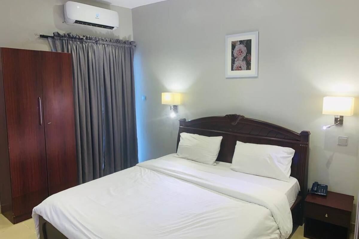 Hotellihuone, jossa on suuri sänky, vaatekaappi, yöpöytä ja seinälle asennettu ilmastointilaite.