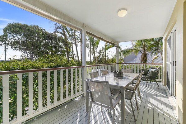 A covered outdoor patio with a table and chairs, surrounded by a white railing and lush greenery.