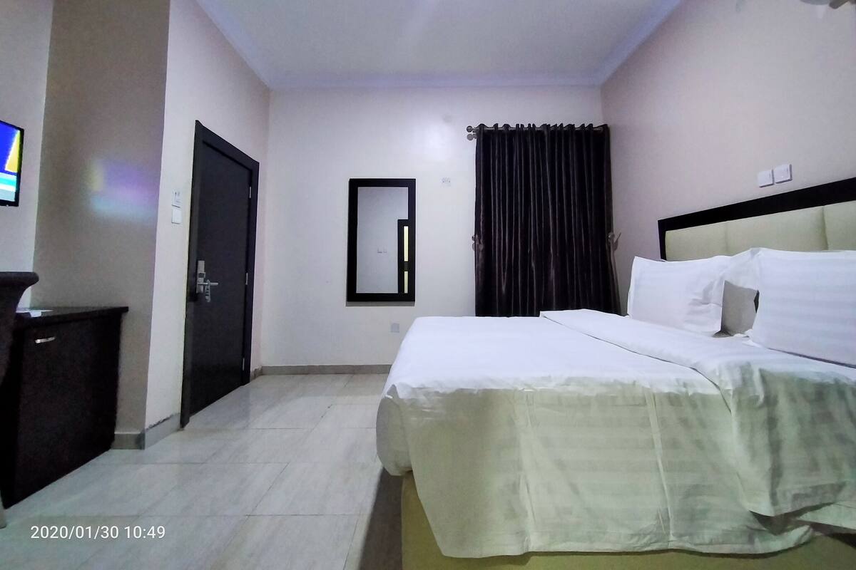 A hotel room with a bed, a TV, a mirror, a window with curtains, and an air conditioning unit.
