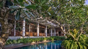 A swimming pool with a wooden deck, surrounded by lush greenery and a covered outdoor area with columns and wooden railings. - The Postcard Galle (Galle)