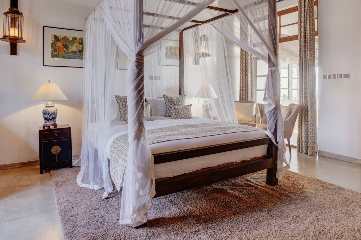 Una cama con dosel sostenido por cuatro pilares, un marco de madera, una mesita de noche con una lámpara, una silla y un cuadro en la pared.
