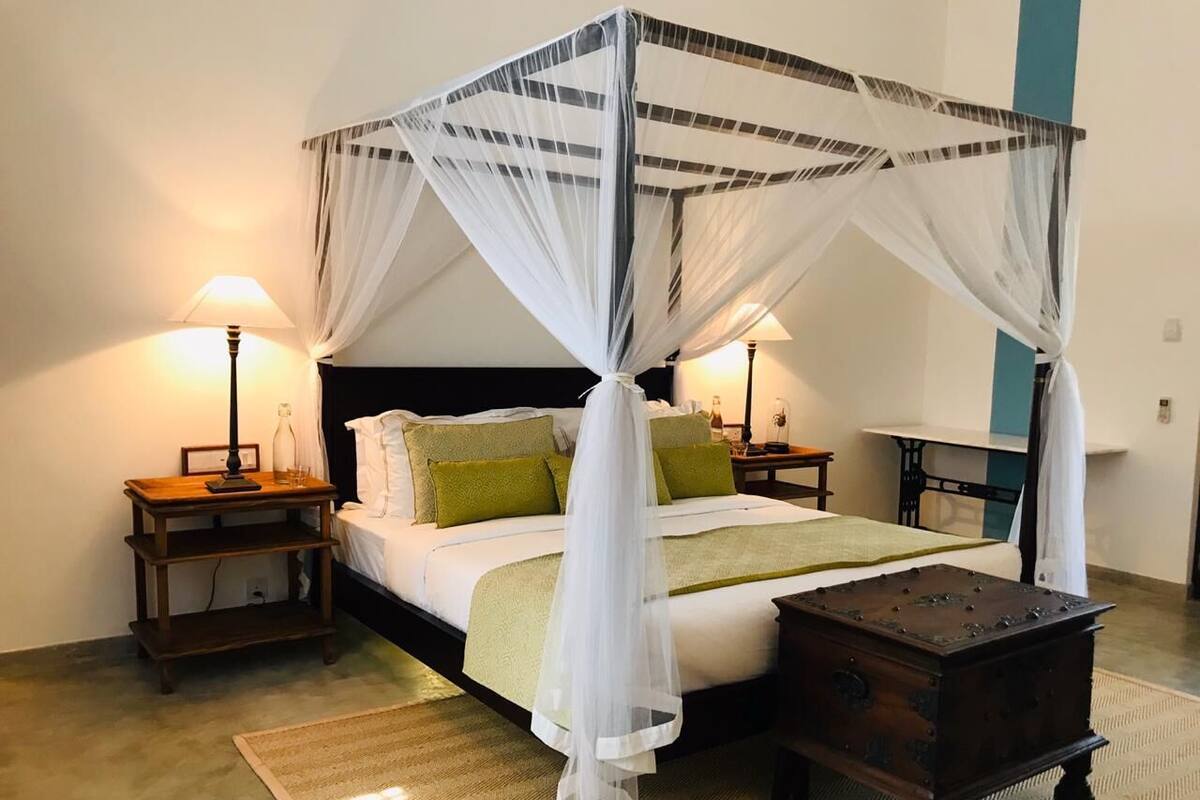 Una cama con dosel y cortinas, ropa de cama blanca, almohadas verdes, dos lámparas de noche, una mesita de madera y un pequeño cofre de madera.