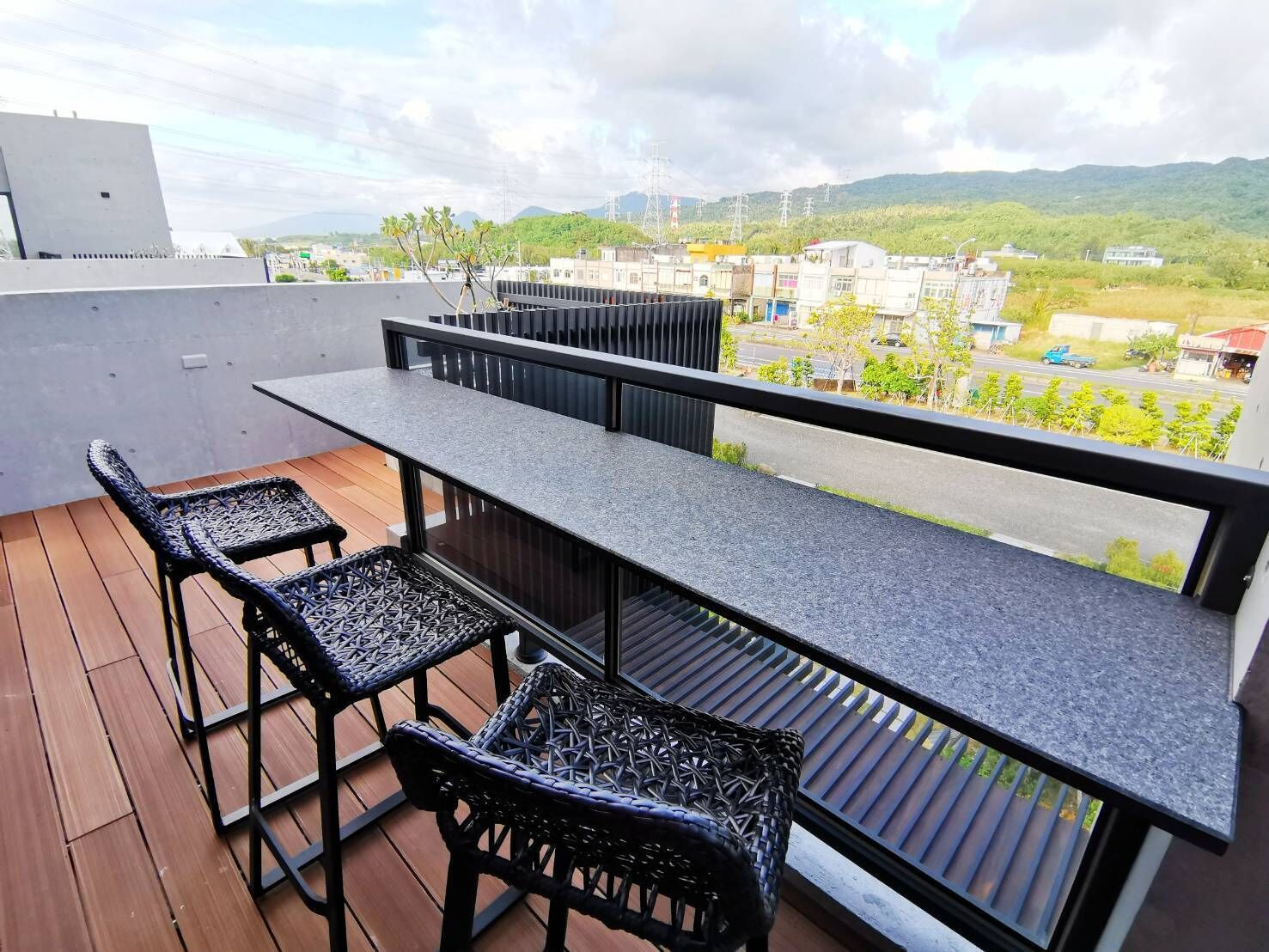 A balcony with a long table and four wicker chairs, overlooking a residential area with buildings and greenery.