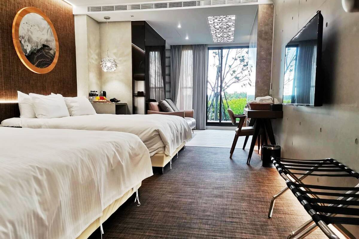 A hotel room with two beds, a desk, a chair, a TV, and a large window with a view of greenery.