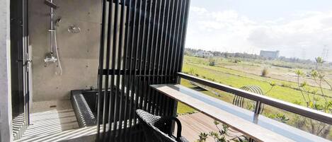 A balcony with a view of a green landscape, a black wicker chair, and a shower area with a vertical privacy screen.