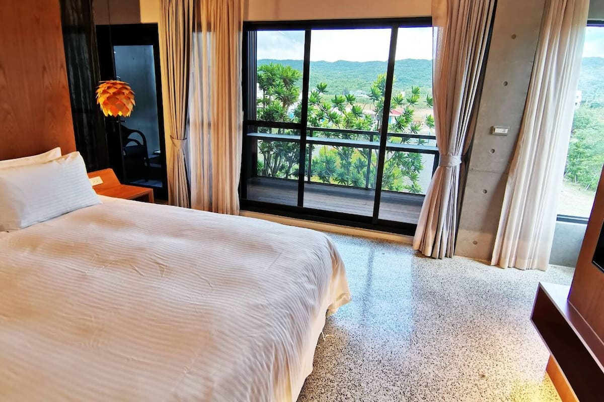 A hotel room with a large bed, a desk, and a view of greenery through a large window.