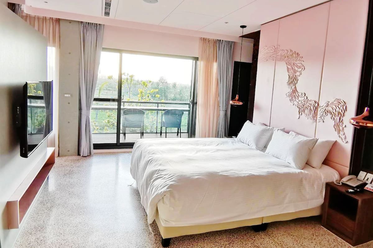 A modern hotel room with a large bed, a television, a balcony with a view, and a decorative wall piece.