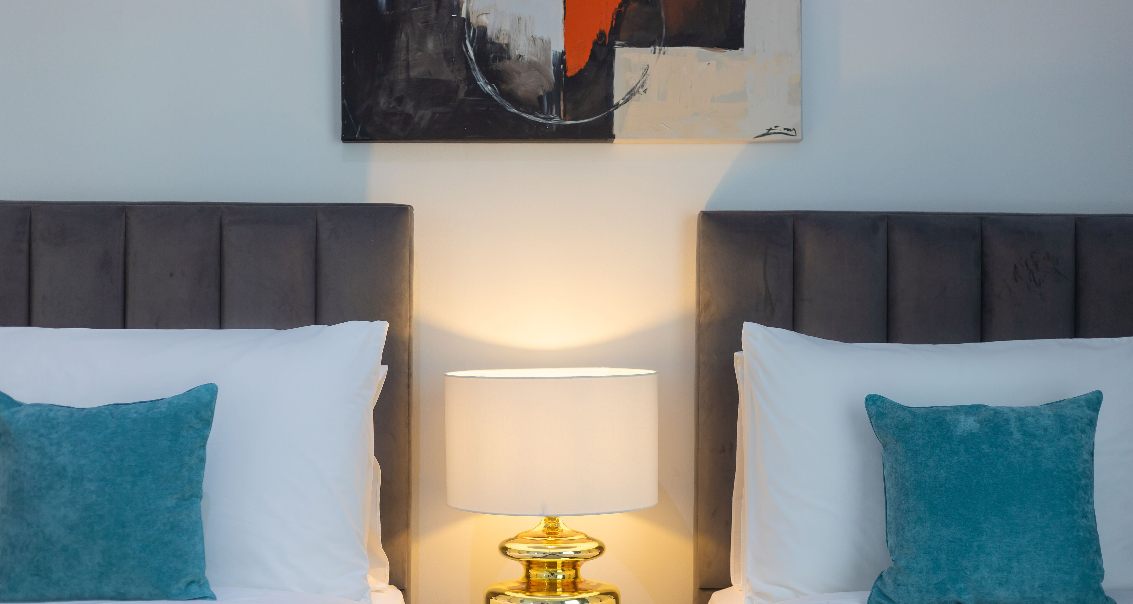 A hotel room with two beds, each with a teal pillow, a white pillow, and a gold lamp between them.