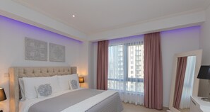 A bedroom with a large bed, two wall art pieces, a window with sheer curtains, and a bedside lamp.