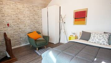 Un dormitorio con pared de piedra, una cama con ropa de cama gris, un sillón verde con un cojín naranja y un armario blanco.