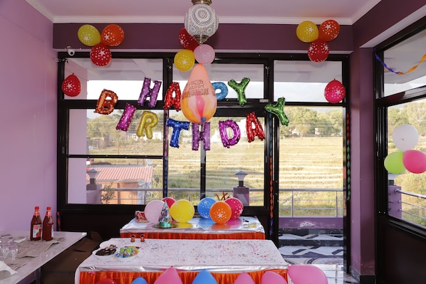 A room with a birthday theme, including balloons, a table with a birthday cake, and a window with a view of a field.