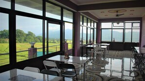 A spacious room with large windows offering a view of a green landscape and mountains. There are several tables and chairs arranged for dining or meetings. - Billing Vista Guest House (Jogindarnagar)