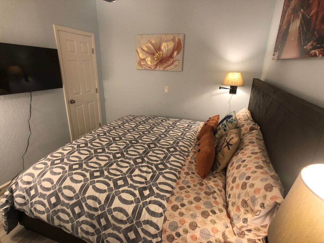 Un dormitorio con una colcha estampada, un televisor montado en la pared, una puerta y una lámpara de pared.