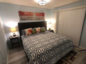 A bedroom with a patterned bedspread, two bedside lamps, a headboard, and a painting on the wall.