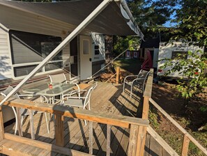 A covered outdoor deck with a table and chairs, a wooden deck, and a camper parked nearby. - Springdale Trailer at Bon Echo Family Campground | Rustic Stay w/ Beach Access
 (Cloyne)