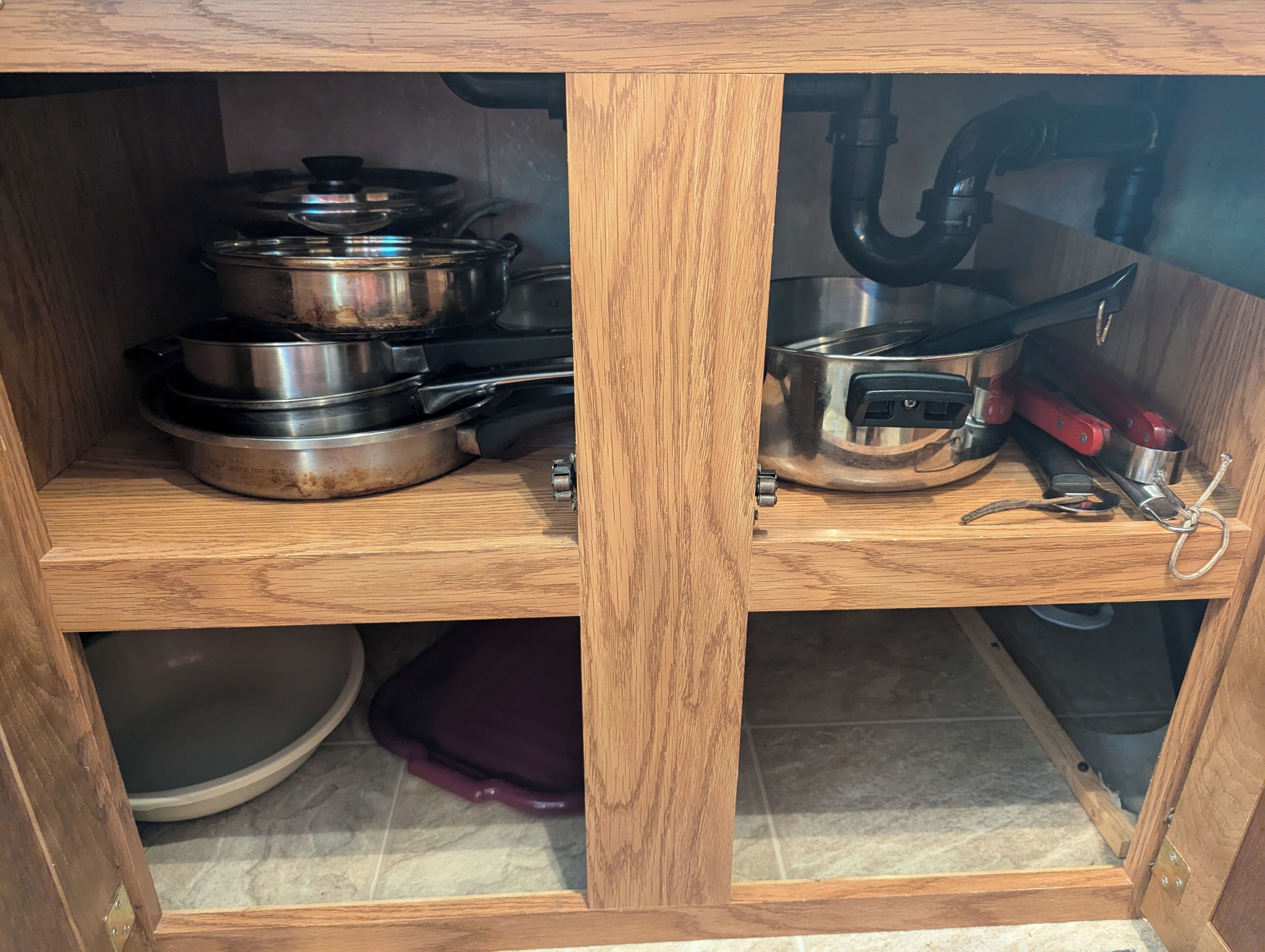 A wooden cabinet with metal cookware, including pots and pans, and a red-handled utensil.
