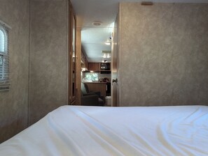 A bedroom with a large bed, a wooden door leading to a kitchenette, and a window with blinds. - Springdale Trailer at Bon Echo Family Campground | Rustic Stay w/ Beach Access
 (Cloyne)