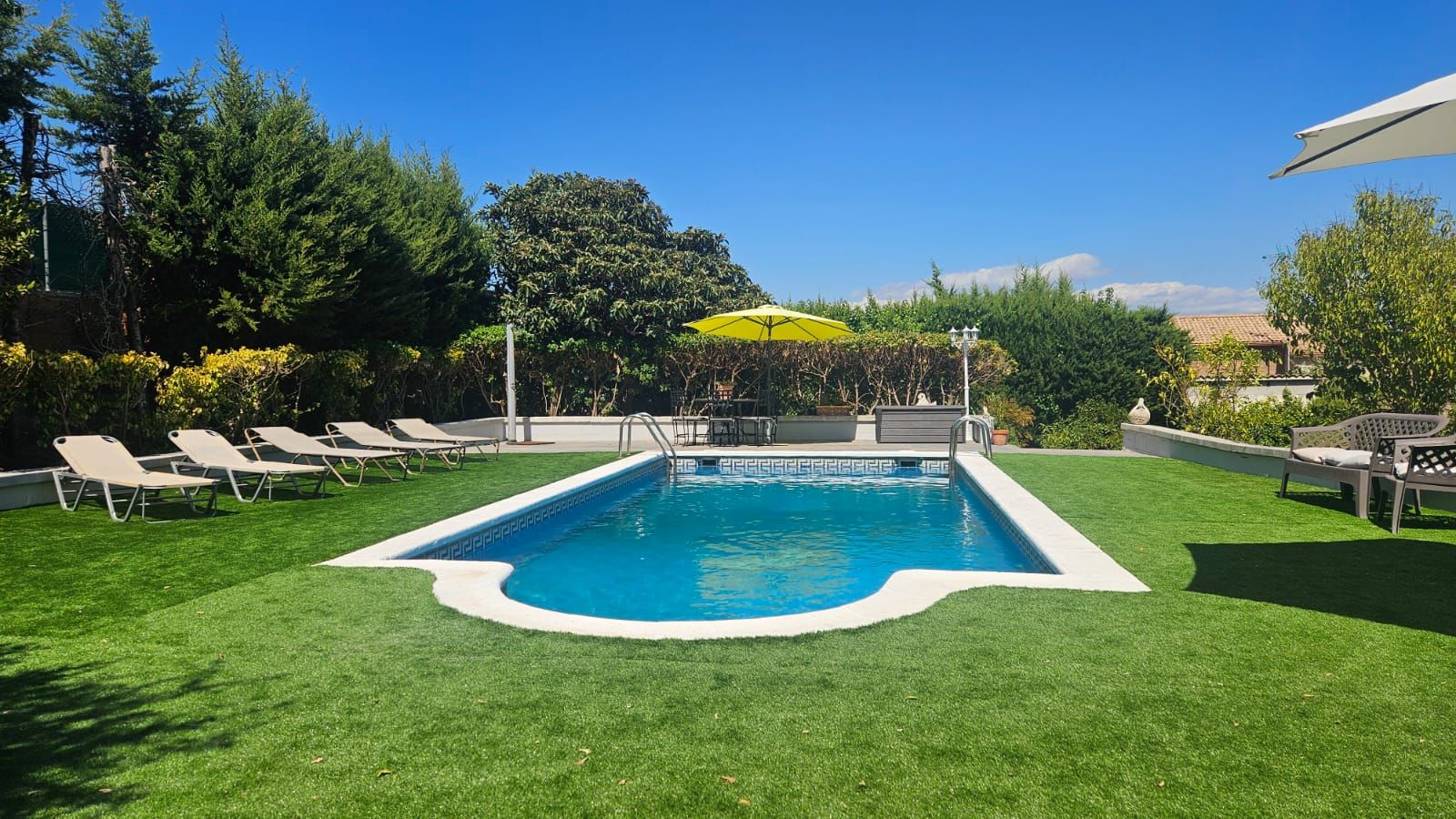 Una piscina rectangular con borde blanco, rodeada de césped cuidado y sillas de descanso.
