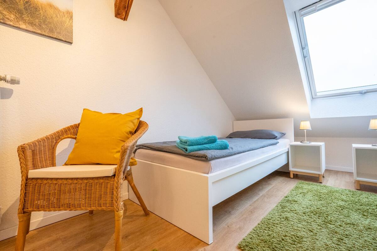 A small, cozy room with a bed, a wicker chair with a yellow cushion, a green rug, and a window providing natural light.