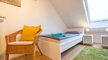 Een kleine, gezellige kamer met een bed, een rieten stoel met een geel kussen, een groene vloerkleed en een raam dat natuurlijk licht biedt.