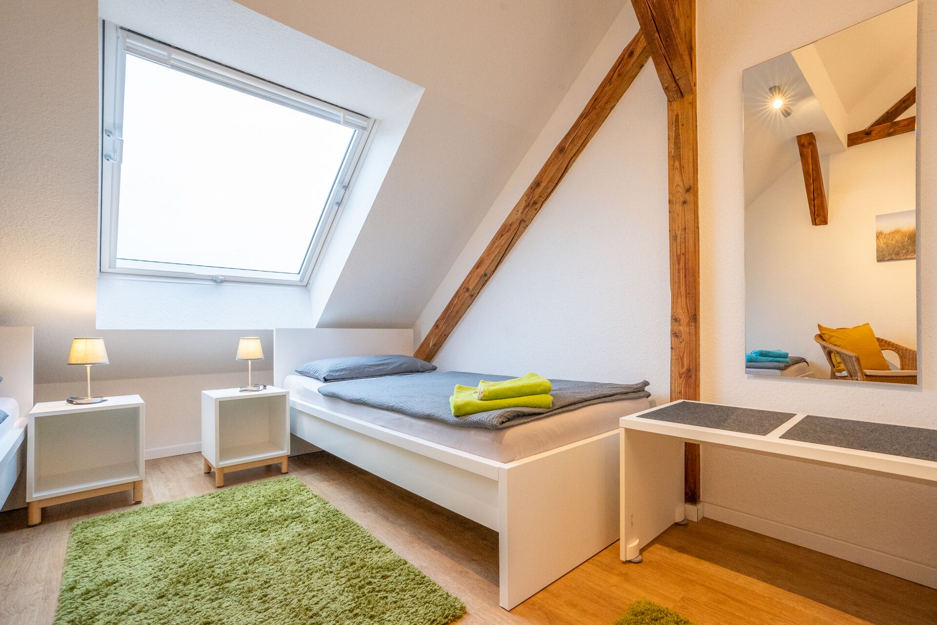 Un dormitorio con techo inclinado, tragaluz, cama con colchón y almohadas azules, mesita de noche con lámpara, alfombra verde y una pequeña zona de escritorio.