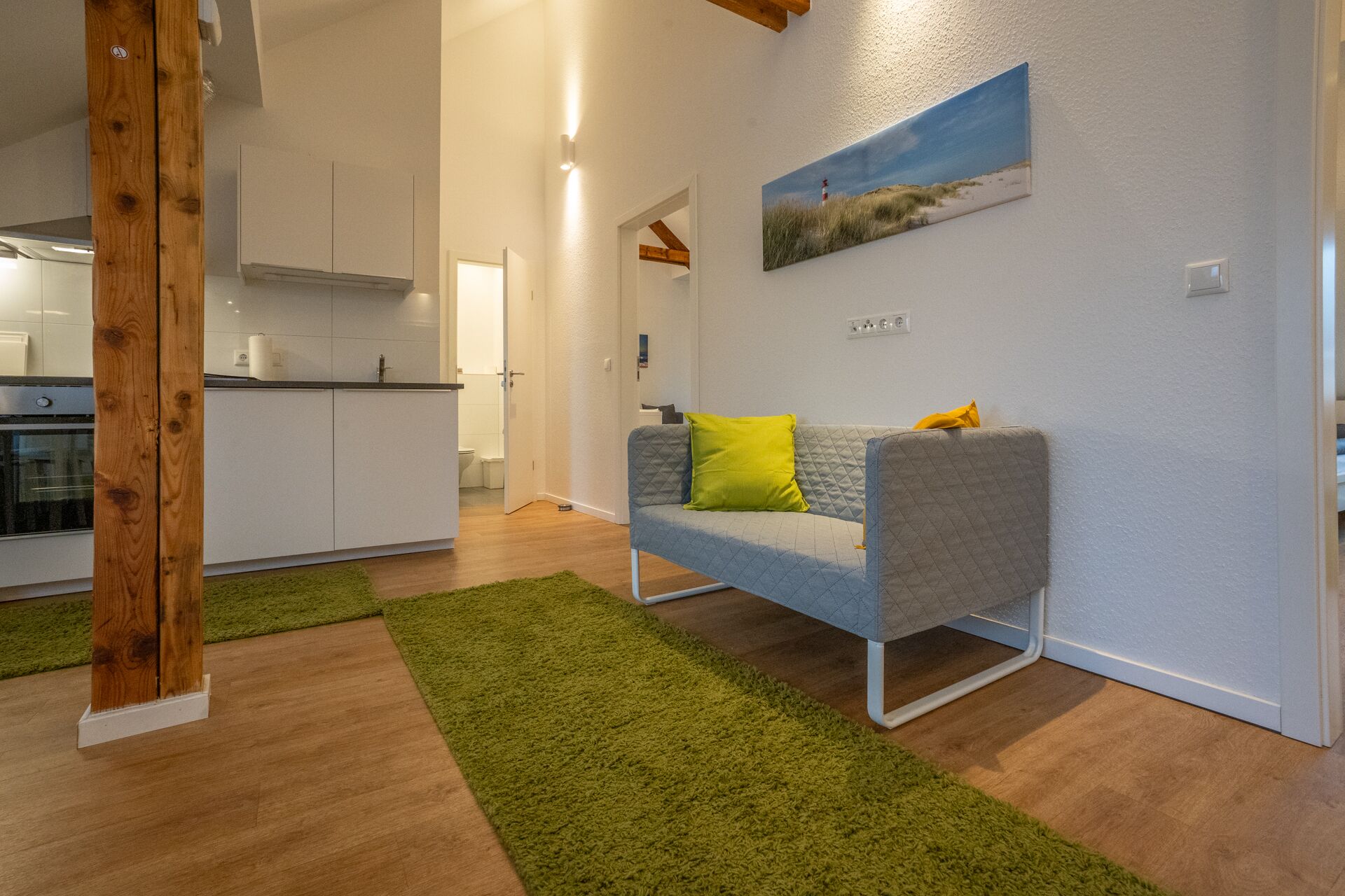 A modern living room with a grey sofa, green and brown rugs, a painting of a beach scene, and a kitchen in the background.