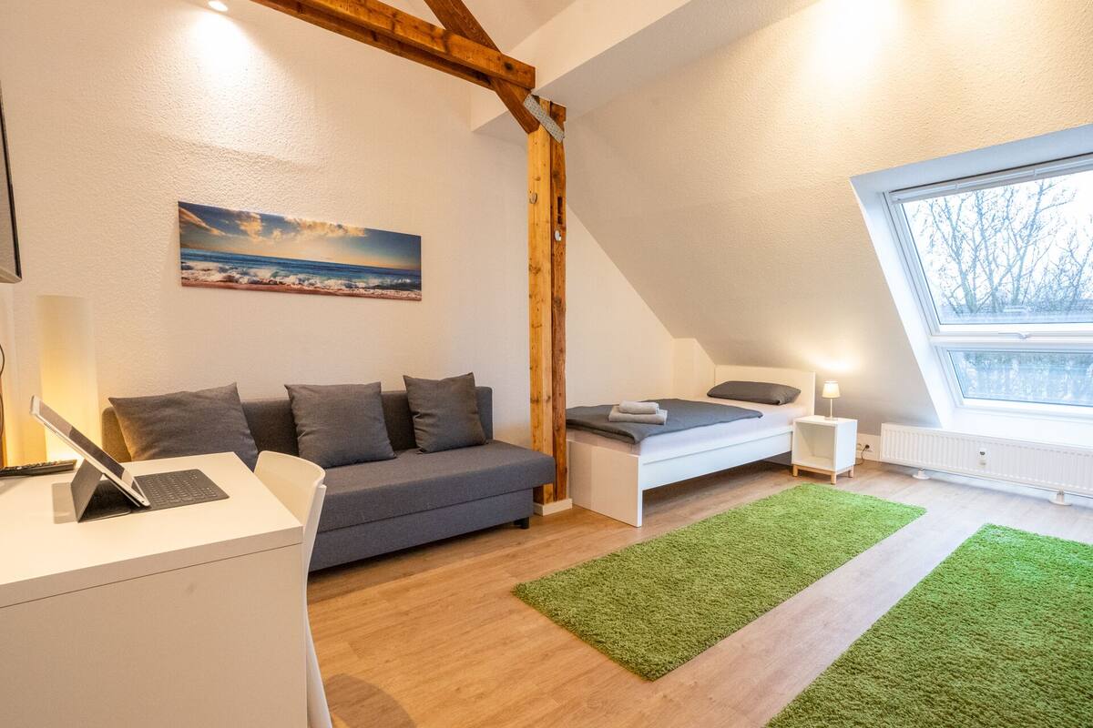 A modern bedroom with a sloped ceiling, a gray sofa, a small bed, a desk with a laptop, and a large window with a view of trees.