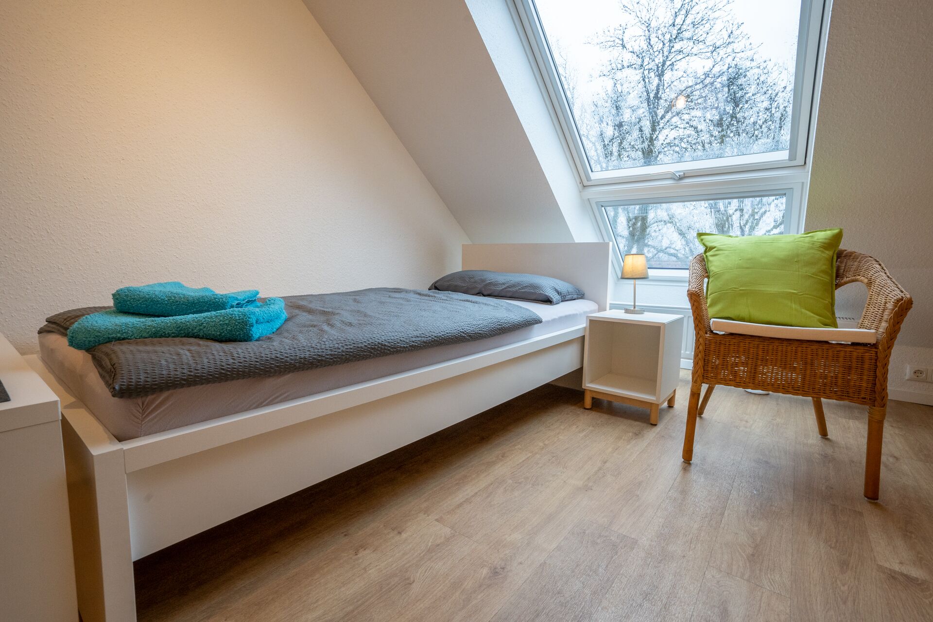 A small, single-bed room with a sloped ceiling, a wicker chair, a bedside table with a lamp, and a window with a view of trees.