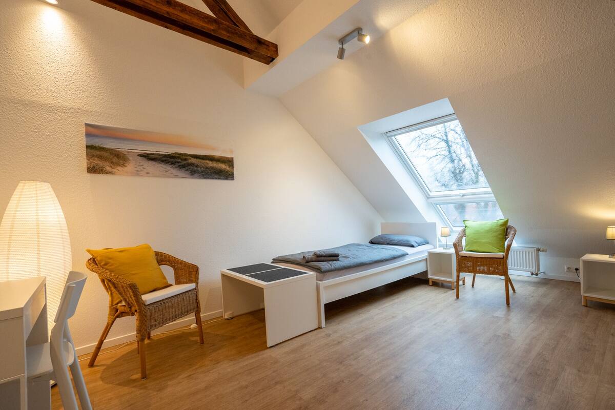 A small, cozy room with a loft bed, a window, a chair, and a lamp.