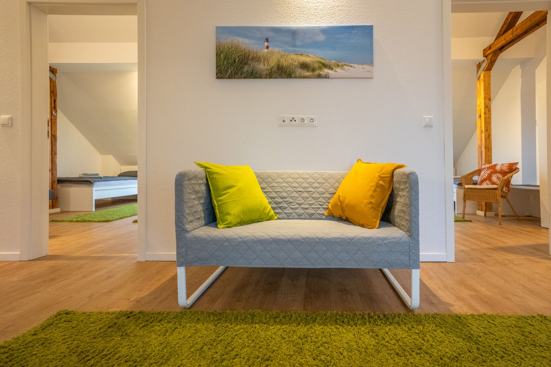 Una sala de estar moderna con un sofá gris, dos cojines amarillo brillante, una alfombra verde y un cuadro de un faro en la pared.