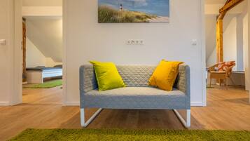 Una sala de estar moderna con un sofá gris, dos cojines amarillo brillante, una alfombra verde y un cuadro de un faro en la pared.