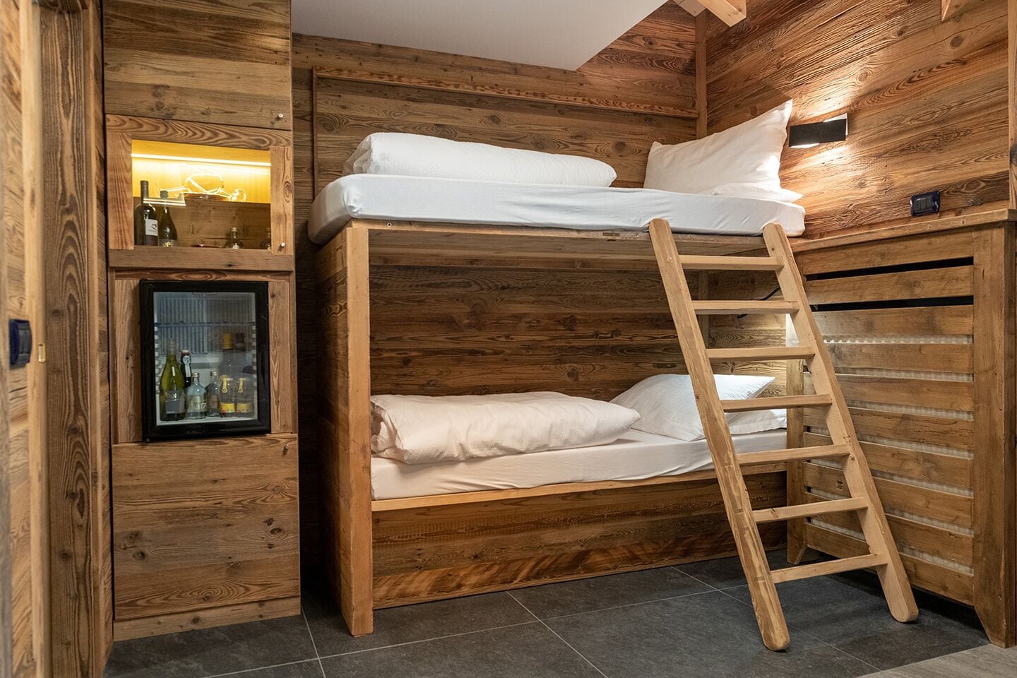 Una cama individual superpuesta con escalera, paredes de madera y una minibar con botellas.