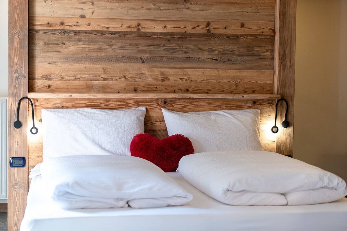 A bed with white linens, two pillows, and a red heart-shaped pillow.