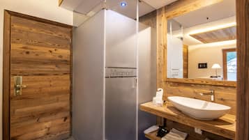 Ein modernes Badezimmer mit Holztür, einer Duschkabine aus Glas mit dem Schriftzug "Miramonti", einem hölzernen Waschtisch mit weißem Waschbecken und einem Spiegel.