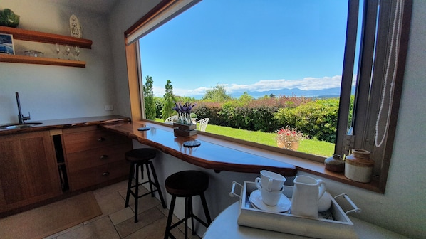 Eine Küche mit großem Fenster, Holztisch, Hockern und Blick auf Garten und Berge.