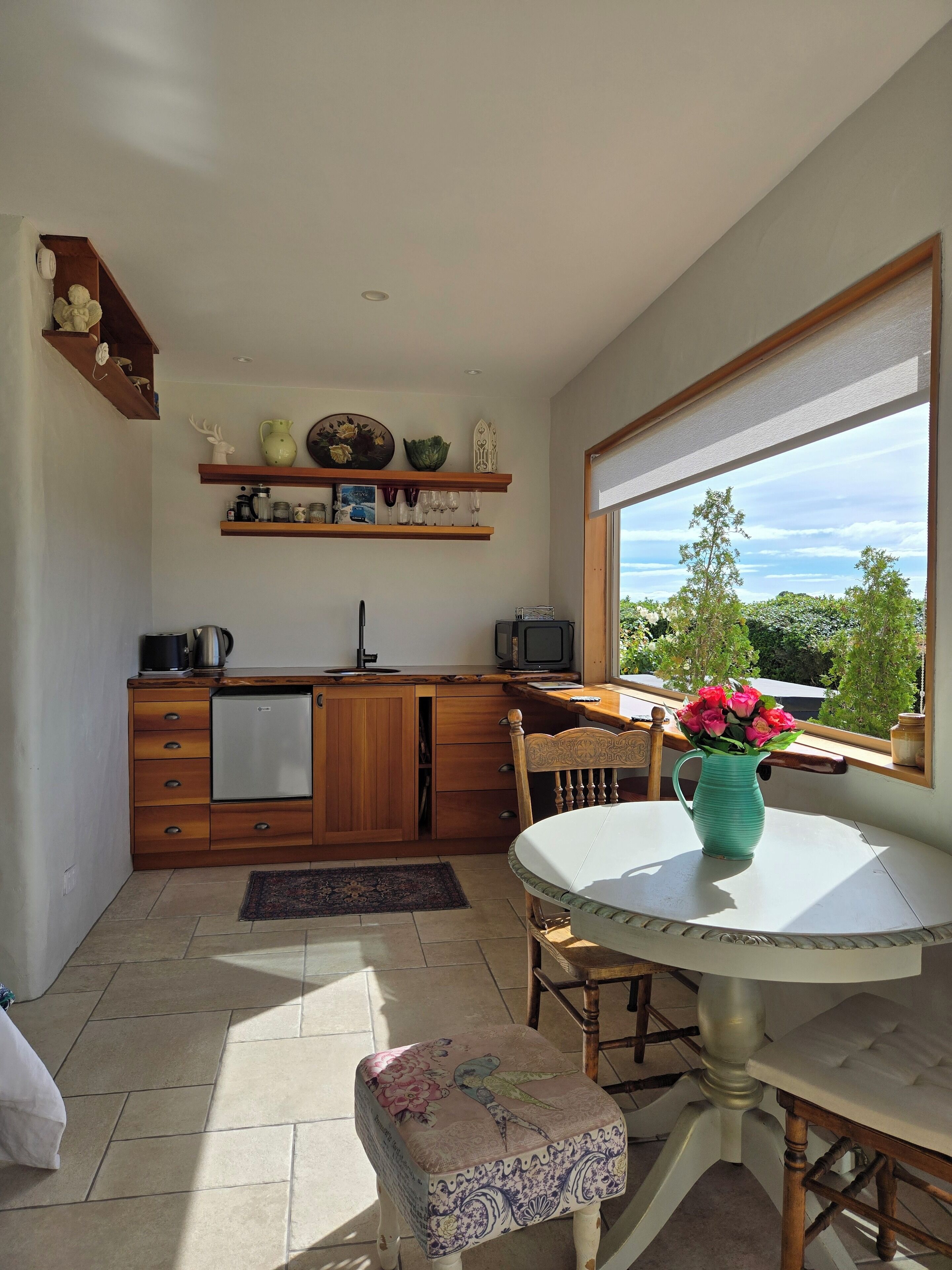 A compact kitchen with wooden cabinets, a dining table with a vase of flowers, and a window overlooking greenery.