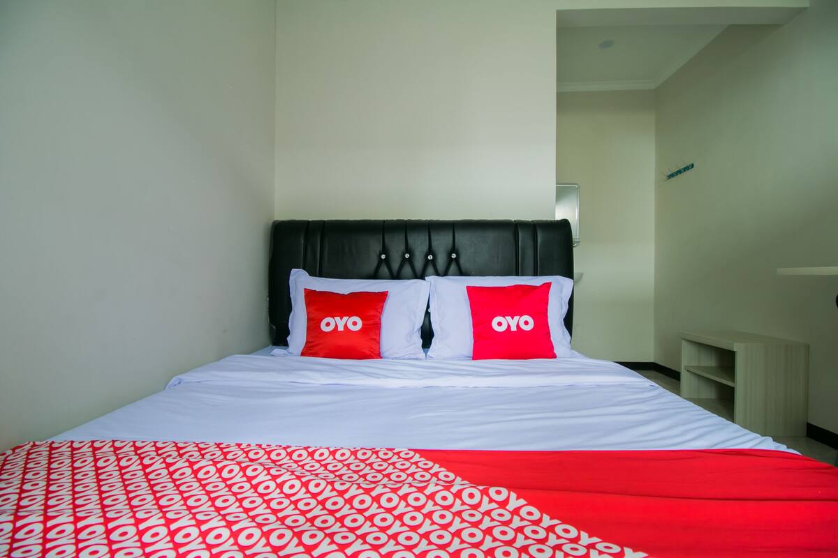 A hotel room with a bed, two red pillows with the word "OYO" on them, a white bedspread, and a red and white patterned blanket.