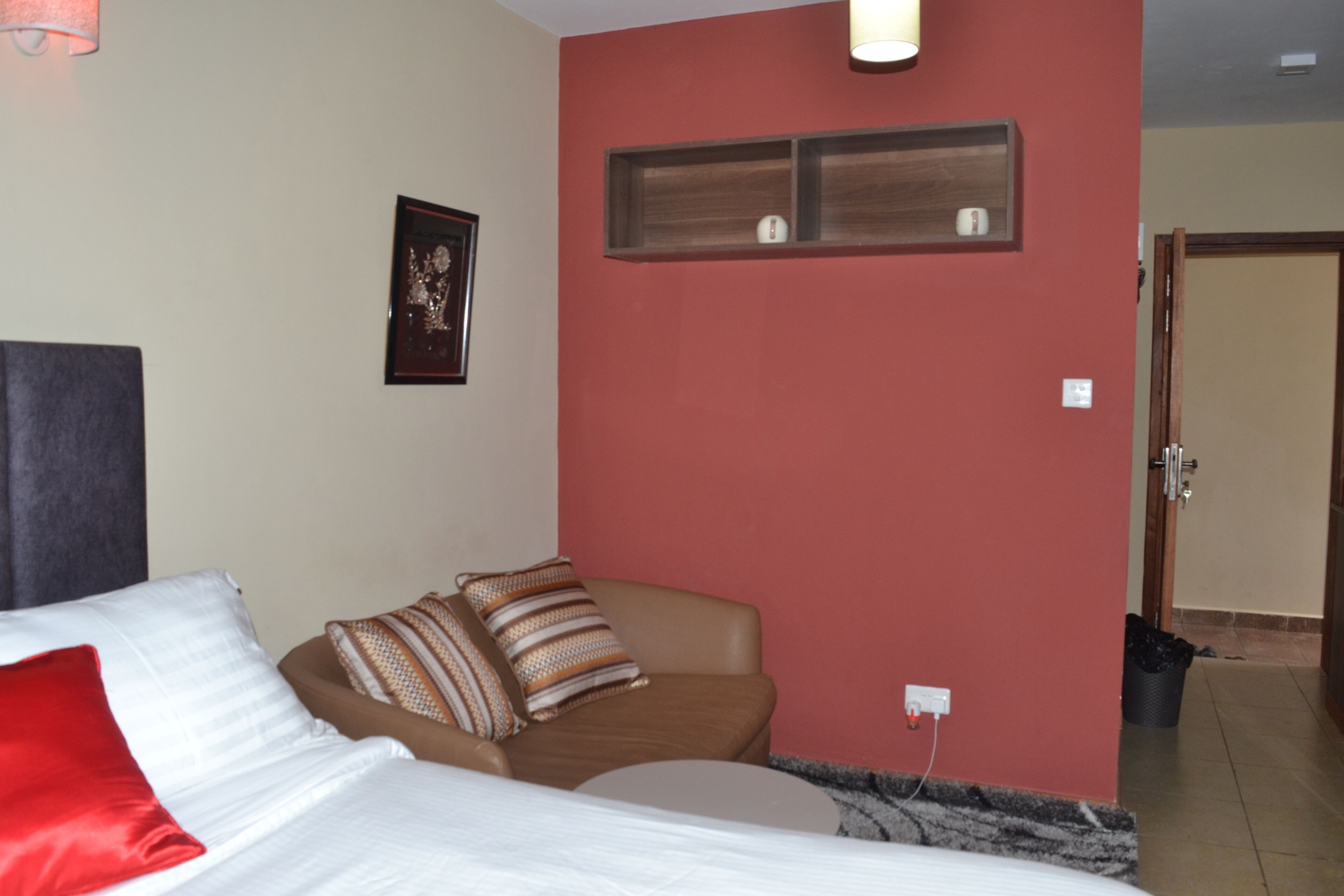 A hotel room with a bed, a chair, a red wall, a framed picture, and a door.